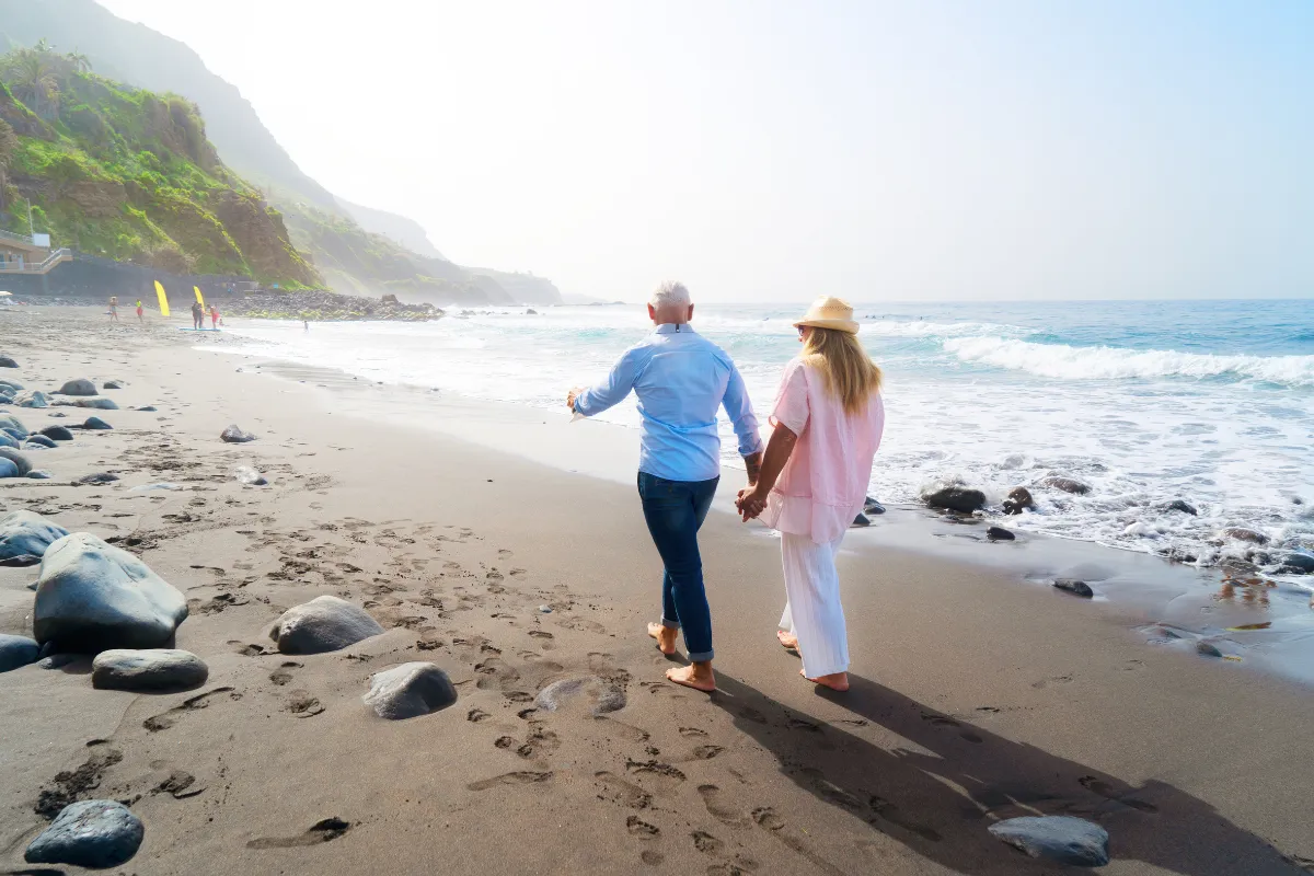 An older couple holding hands while walking along a rocky beach
