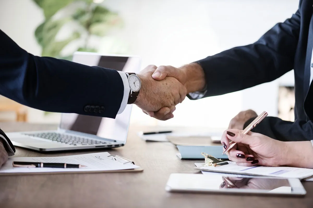 Two business people in suits shake hands over a desk with a laptop and paperwork