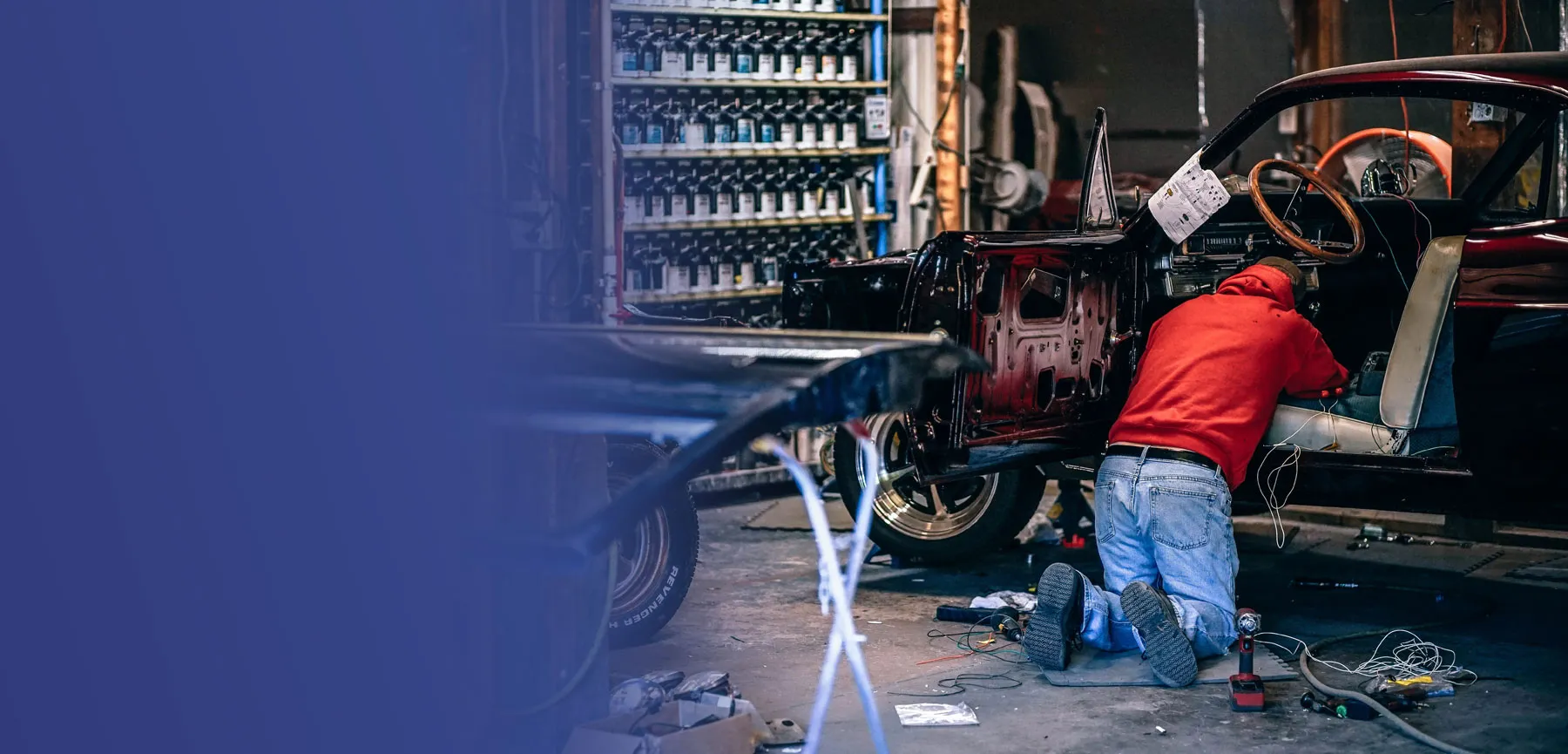 A mechanic in a red shirt works on the stripped-down interior of a classic car in a garage